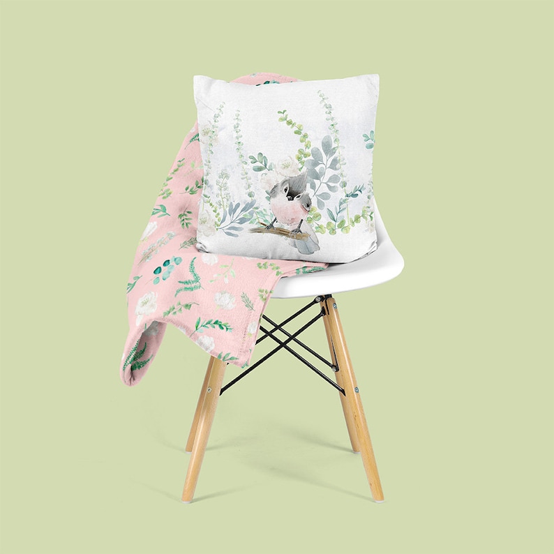 May include: A pink throw blanket with a floral pattern featuring white flowers and green leaves is draped over a white chair. A white pillow with a watercolor illustration of a bird perched on a branch with green leaves is on the chair.