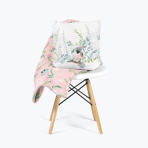 May include: A pink throw blanket with a floral pattern featuring white flowers and green leaves is draped over a white chair. A white pillow with a watercolor illustration of a bird sitting on a branch with green leaves is on top of the blanket.