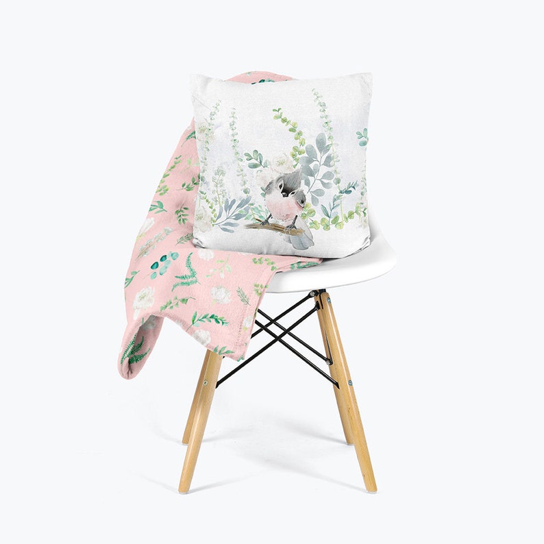 May include: A pink throw blanket with a floral pattern featuring white flowers and green leaves is draped over a white chair. A white pillow with a watercolor illustration of a bird sitting on a branch with green leaves is on top of the blanket.