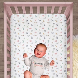 May include: A white crib sheet featuring a repeating pattern of small, multi-colored cat silhouettes in shades of pink, blue, green, and orange. The sheet is inside a light pink wooden crib, with a baby wearing a long-sleeve gray bodysuit.