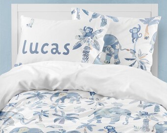 May include: White bedding with a blue and white jungle print featuring elephants, monkeys, and palm trees. The pillowcase has the name "lucas" written in blue.