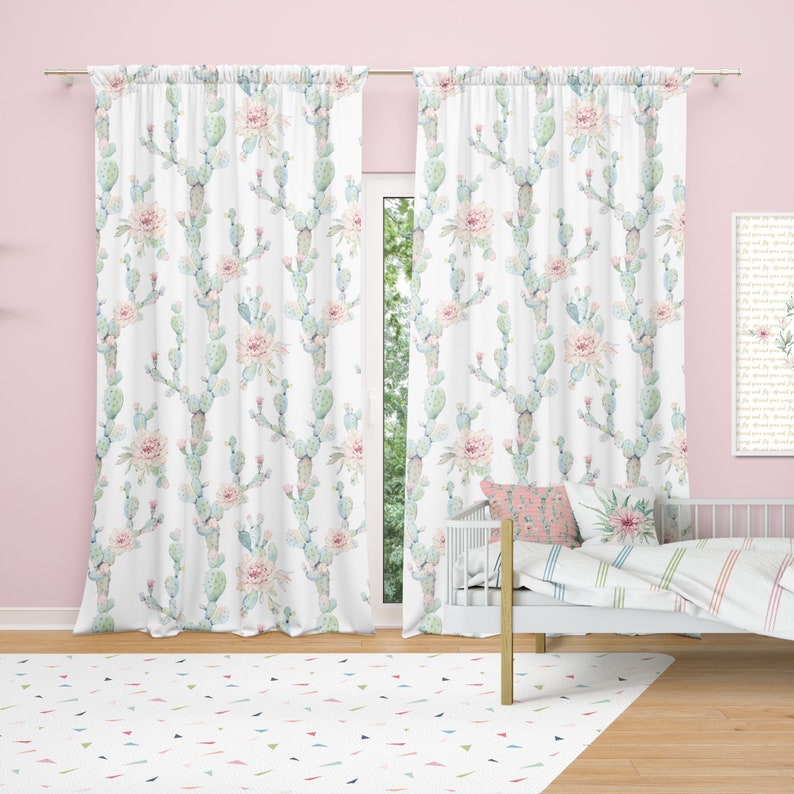 May include: White curtains with a repeating pattern of light blue cacti and pink flowers. The curtains hang in front of a window, and the room has a pink wall and a white bed with a patterned pillow.