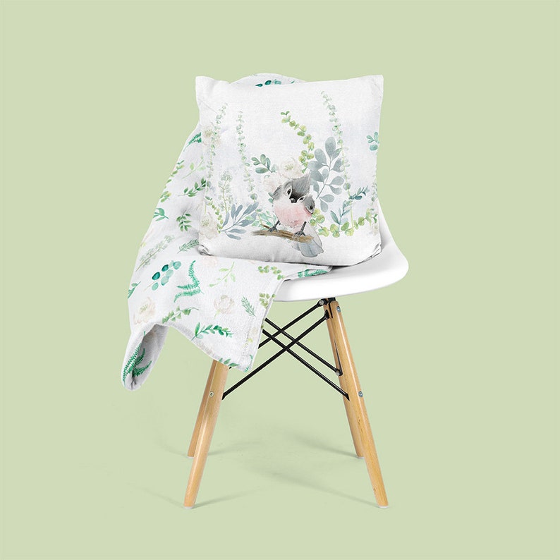 May include: A white chair with a white and green floral patterned throw blanket and a white pillow with a watercolor painting of a bird on a branch. The chair is on a light green background.