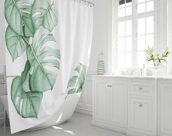 Pode incluir: Uma cortina de duche branca com uma grande ilustração em estilo aguarela de folhas de monstera verdes. O design está posicionado no lado esquerdo da cortina, que está pendurada numa barra com argolas visíveis, cobrindo a área da banheira.