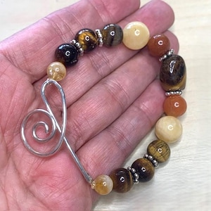 Op de afbeelding: Een armband met een mix van bruine, gele en oranje edelstenen. Het heeft een zilverkleurige, gestileerde muzieksleutelbedel. Kleine zilveren afstandhouders zitten tussen de kralen.