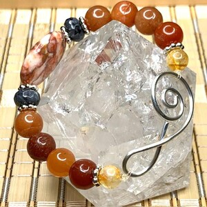 Op de afbeelding: Een kralenarmband met oranje, rode en gele edelsteen kralen. De armband heeft een ovale kraal met een patroon en een zilveren spiraalvorm. De kralen zijn geregen met zilverkleurig draad.