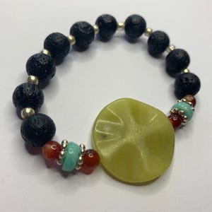Op de afbeelding: Een elastische armband met zwarte poreuze lavasteenkralen, kleine zilverkleurige tussenstukjes, gladde roodbruine carneoolkralen, lichtblauwe turkooiskleurige schijfjes en een grote, platte, limoengroene centrale kraal met een kruispatroon.