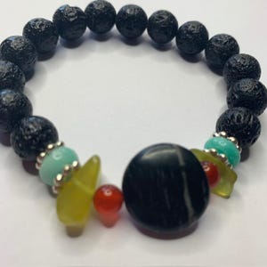 Op de afbeelding: Een elastische armband met een reeks ronde, poreuze zwarte lavakralen. Het ontwerp bevat een grote, gladde zwarte ronde centrale kraal, geaccentueerd door kleinere ronde rode kralen, doorschijnende geelgroene scherven en lichtblauwe kralen.