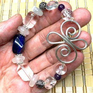Op de afbeelding: Een kralenarmband met heldere, witte en blauwe edelstenen, decoratieve zilveren kralen en een groot zilveren spiraalontwerp. De armband wordt in een hand getoond, waarbij de kleuren en texturen worden getoond.