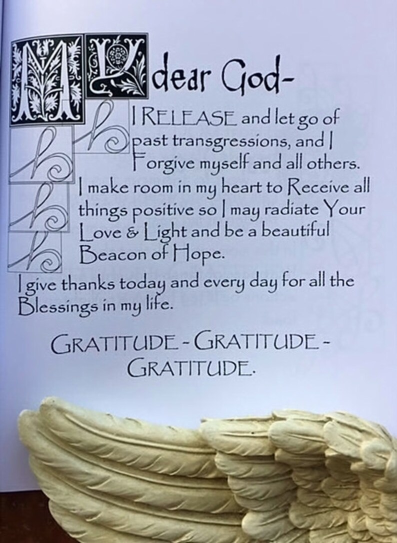 May include: A page with handwritten text, including the phrases "dear God," "I RELEASE," and "GRATITUDE." Decorative initial letters and a sculpted wing are visible. The text is a prayer or affirmation.
