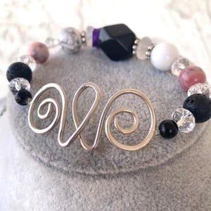 Op de afbeelding: Een armband met een zilveren draadontwerp en diverse kralen. De kralen zijn zwart, wit, roze en helder, met een grote zwarte geometrische kraal. De armband wordt gepresenteerd op een grijze ondergrond.