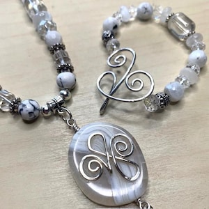 Op de afbeelding: Een handgemaakte ketting- en armbandset. De ketting heeft witte en heldere kralen, zilveren accenten en een ovale hanger met een zilveren draadontwerp. De armband heeft vergelijkbare kralen. De kralen zijn in verschillende maten.