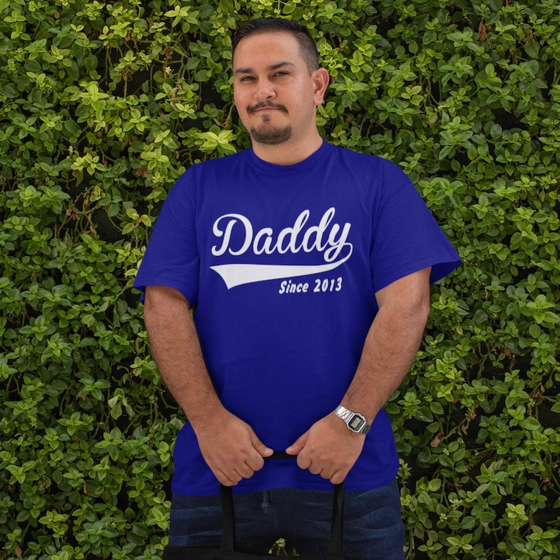 以下が含まれることがあります： ヴィンテージの野球ユニフォーム風のフォントで「Daddy Since 2013」と書かれた白いテキストが入ったロイヤルブルーのTシャツです。