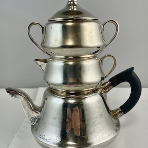 May include: A silver three-tiered teapot with a black handle. The top tier is a sugar bowl with a lid. The teapot has a rounded body and a spout.