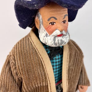 May include: A handmade doll with a white beard and mustache, wearing a brown corduroy jacket, a blue and white plaid shirt, and a dark blue hat.