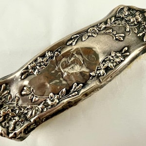 May include: An elongated silver hairbrush with a polished metallic finish. The handle and edges feature intricate embossed floral and leaf patterns. A central smooth panel displays an engraved monogram with cursive lettering in the middle.