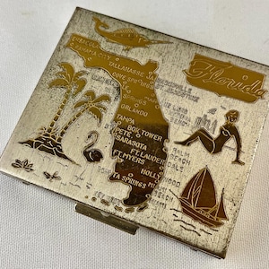 May include: A silver compact with a gold-toned design of a map of Florida with various cities and landmarks labeled. The compact has a hinged lid and a mirror inside. The word "Florida" is written in gold letters at the top of the compact.