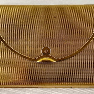 May include: A rectangular, gold-toned metal case with a textured, cross-hatched pattern. The case features a curved flap with a small, circular clasp. The overall design is vintage and elegant.