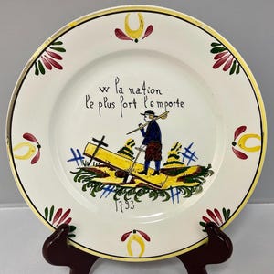 May include: A white ceramic plate with a yellow rim and decorative floral accents. The plate features a hand-painted scene of a person carrying a yellow object, with the text "w la nation le plus fort l'emporte" and the year "1793".