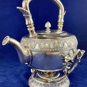 May include: A silver teapot with an ornate design and a stand. The teapot has a curved handle and a spout. The stand has three legs and a round base.