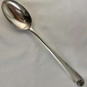 May include: A shiny, silver spoon with a long, slender handle and an oval bowl. The spoon reflects the surrounding environment, creating a mirrored effect. The spoon is likely made of stainless steel or a similar metal.