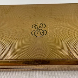 May include: A gold-colored metal compact with a circular design and the initials "GS" engraved in the center.