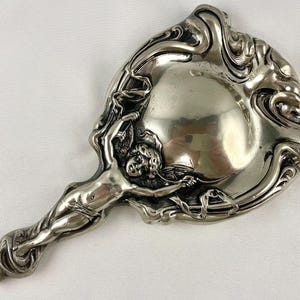 May include: A silver-toned hand mirror featuring an ornate, embossed frame with swirling, organic patterns. The handle and base of the mirror are integrated into a sculptural relief of a winged cherub figure with detailed facial features.