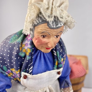 May include: A vintage doll of an elderly woman wearing a white bonnet, a blue dress with a floral print, and a white apron. The doll is sitting down and has a realistic face.