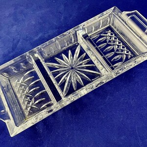 May include: A clear, rectangular glass serving tray with three compartments and two handles. The tray features intricate etched patterns, including a starburst design in the center and diamond and leaf patterns in the side sections. The tray is ideal for serving snacks or appetizers.