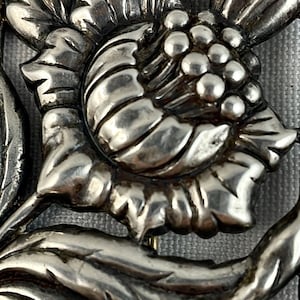 May include: A silver floral brooch with a detailed design of a flower with a cluster of round buds in the center.