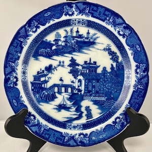 May include: A blue and white porcelain plate with a scalloped edge. The plate features a detailed landscape scene with buildings, trees, and a boat. The border has a repeating pattern. The plate is displayed on a black stand.