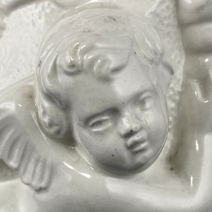May include: White ceramic sculpture of an angel with wings, a cherub face, and curly hair. The angel is depicted with its arms crossed and its head tilted slightly to the side.