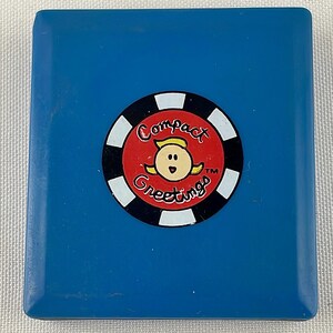 May include: A blue, rectangular compact with a circular emblem. The emblem features a cartoon woman with blonde hair, surrounded by the words "Compact Greetings" in a red circle with a black and white border.