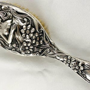 May include: A silver-toned hairbrush featuring an embossed design of a figure with long flowing hair surrounded by floral motifs. The handle and back of the brush are decorated with intricate raised flower patterns and swirling lines, with light-colored bristles visible along the edge.