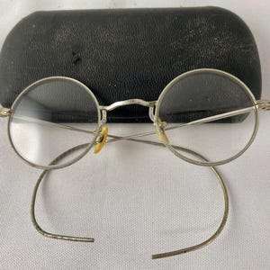 May include: Silver-toned round eyeglasses with a black leather case. The glasses have a simple design with thin metal frames and clear lenses.