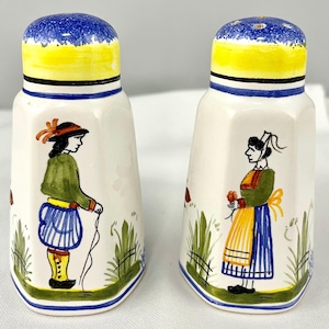 May include: A pair of white ceramic salt and pepper shakers with a hexagonal shape. Each shaker features a hand-painted figure in traditional attire, green grass, and a blue, yellow, and white cap. The shakers have a smooth, glossy finish.