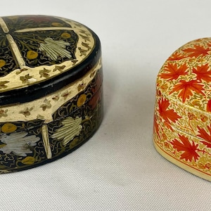 May include: Two decorative boxes. One is oval-shaped with a black base, floral design, and a cream-colored border. The other is square with a red maple leaf pattern on a cream background. Both are closed.