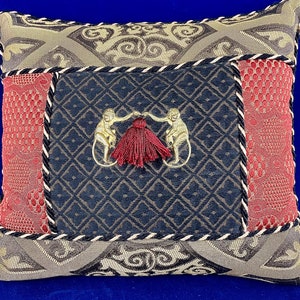 May include: A decorative pillow with a black background and a gold embroidered design of two monkeys holding a red tassel. The pillow is surrounded by a red and gold patterned border.