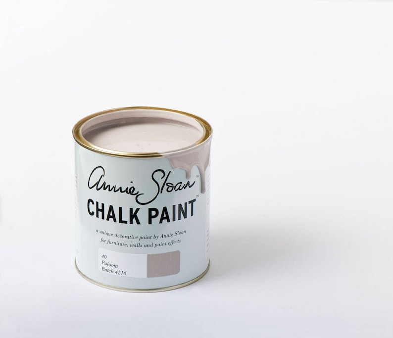 May include: A can of Annie Sloan Chalk Paint in the color Paloma. The can is light blue with gold trim and features the brand name and product information. The paint is a light, neutral color, perfect for furniture and wall projects.