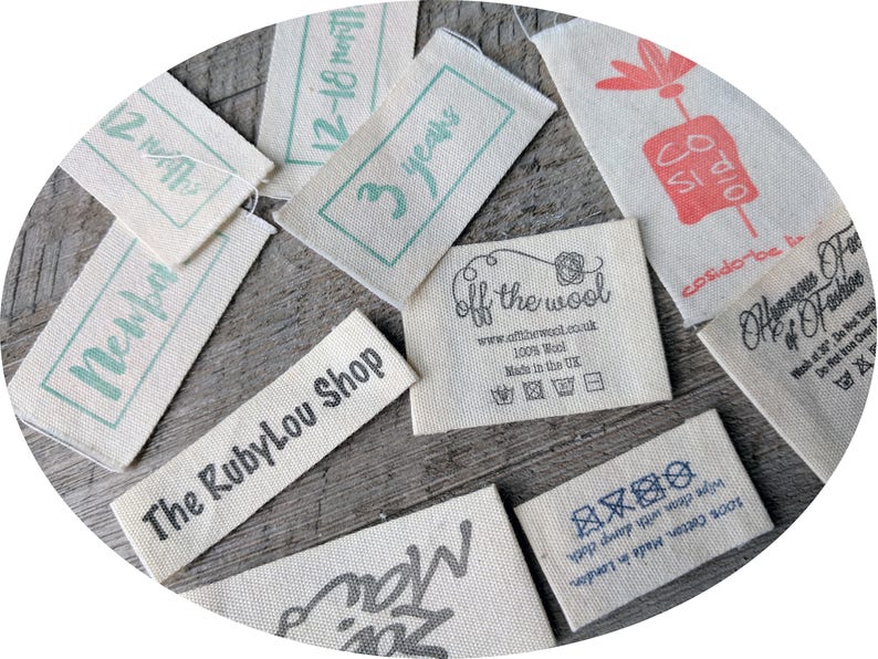 May include: A collection of fabric labels with various text and symbols. The labels are beige with black and green text. The text includes "The RubyLou Shop", "3 years", "off the wool", "12-18 months", "Petal", and "cosido be tin".