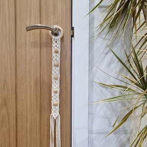 May include: A white macrame door hanger with wooden beads. The hanger is attached to a door handle with a ring.