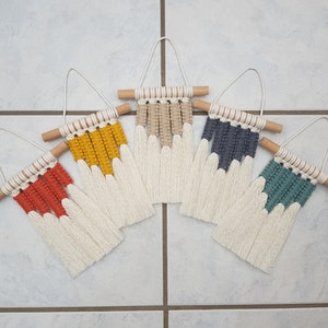 May include: Five macrame wall hangings in rust, yellow, beige, navy, and teal. Each has a wooden dowel, woven top, and fringed bottom. The hangings are displayed on a tiled surface. The measurements are in centimetres.