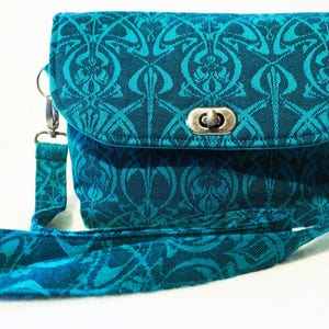 May include: A teal crossbody bag with a flap closure and a silver clasp. The bag and strap feature a repeating floral pattern in shades of teal. The bag is designed with a rectangular shape and a long, adjustable strap.