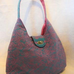 May include: A teal and rose-colored shoulder bag with a rose pattern. The bag has a teal handle with a pink interior, a teal flap with a gold-colored clasp, and a pink interior lining.