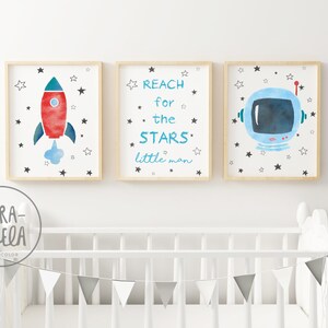 Pu&ograve; includere: Tre stampe ad acquerello incorniciate con un tema spaziale. La prima stampa presenta un razzo rosso e blu con una nuvola di fumo bianca. La seconda stampa ha il testo "REACH for the STARS little man". La terza stampa presenta un casco da astronauta blu con una visiera nera.