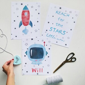 Pu&ograve; includere: Tre poster stampabili a tema spaziale. Il primo poster presenta un razzo rosso e blu con una nuvola bianca dietro. Il secondo poster ha il testo "REACH for the STARS little man". Il terzo poster presenta un casco da astronauta blu con una visiera bianca. Tutti e tre i poster hanno uno sfondo bianco con stelle nere.