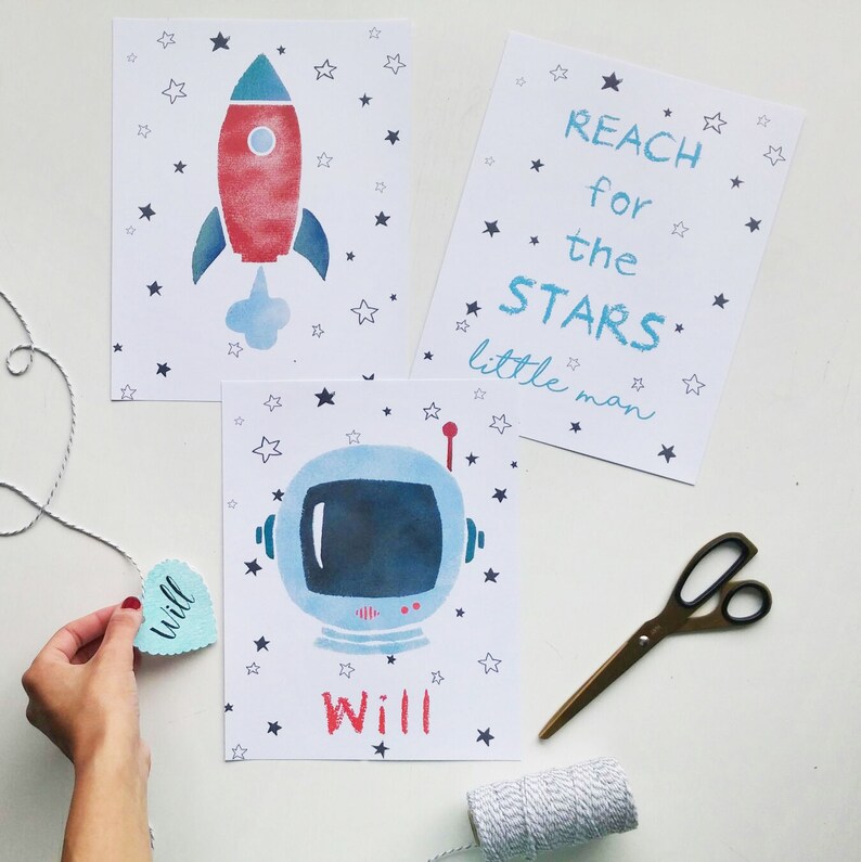 Pu&ograve; includere: Tre poster stampabili a tema spaziale. Il primo poster presenta un razzo rosso e blu con una nuvola bianca dietro. Il secondo poster ha il testo "REACH for the STARS little man". Il terzo poster presenta un casco da astronauta blu con una visiera bianca. Tutti e tre i poster hanno uno sfondo bianco con stelle nere.