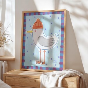 May include: A framed art print featuring a whimsical illustration of a seagull wearing an orange knit cap. The seagull is depicted in white and gray with orange legs and a yellow beak, set against a light blue background with blue polka dots.