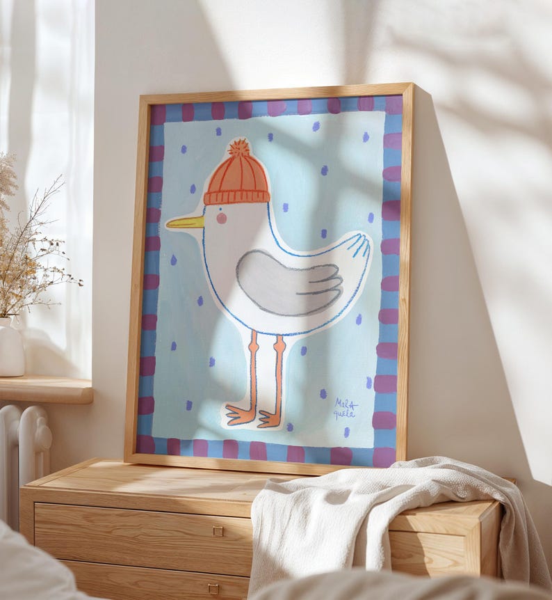 May include: A framed art print featuring a whimsical illustration of a seagull wearing an orange knit cap. The seagull is depicted in white and gray with orange legs and a yellow beak, set against a light blue background with blue polka dots.
