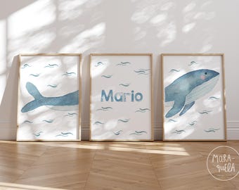 May include: Three framed watercolor prints with a blue whale design. The middle print has the name "Mario" written in blue. The prints are on a light wood floor.